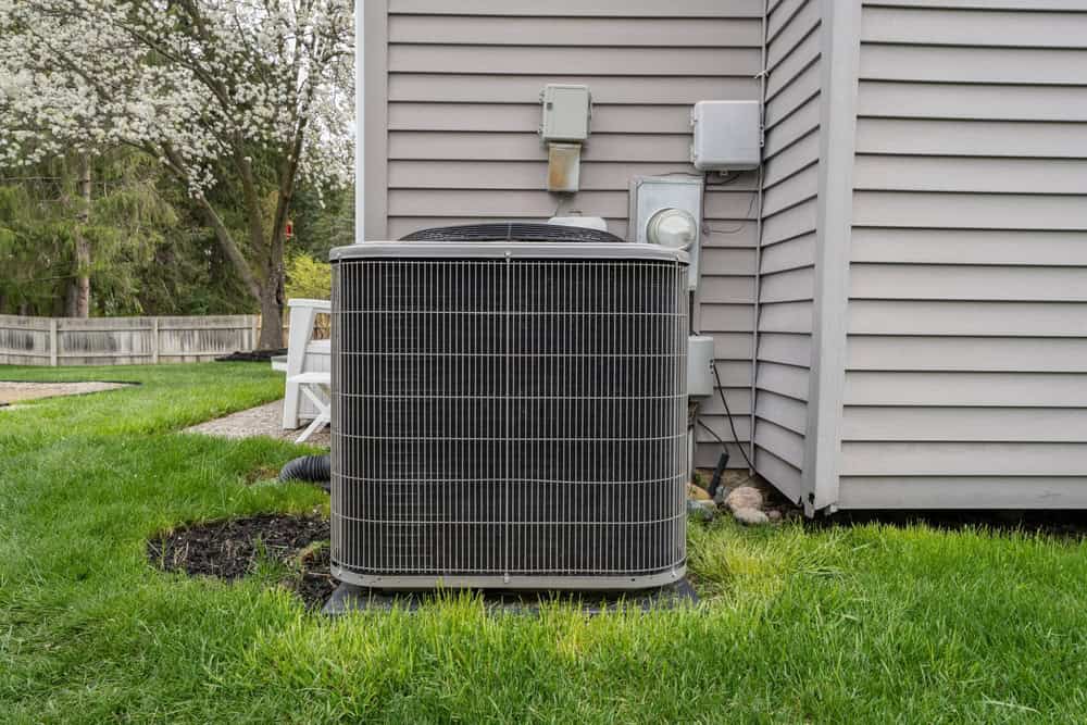 A central air conditioning unit, installed by a trusted HVAC contractor in San Mateo County, CA, sits on a concrete pad outside a house with beige vinyl siding, surrounded by green grass and a flowering tree in the background.