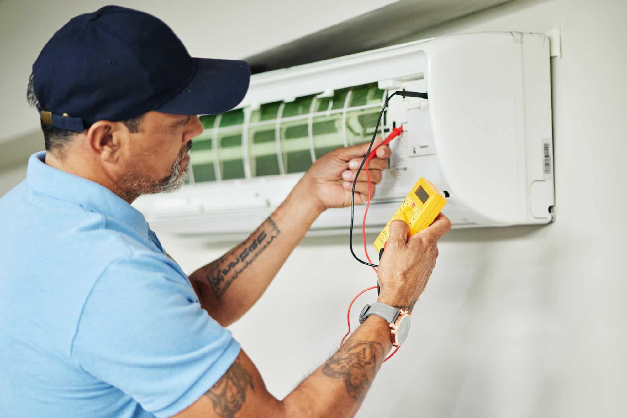 A technician wearing a blue shirt and cap, working for an HVAC contractor in San Mateo County, CA, uses a multimeter to check the electrical components of a wall-mounted air conditioner unit with the front panel open.