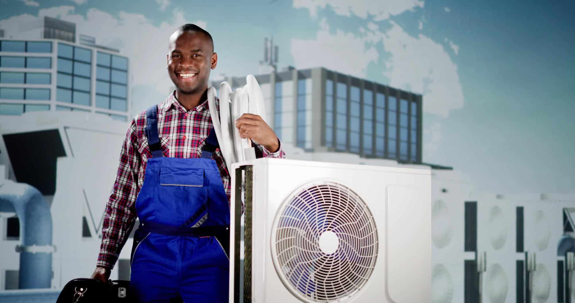 A smiling technician in blue overalls and a plaid shirt stands next to an air conditioning unit, holding tools and a white pipe, proudly representing your trusted HVAC contractor San Mateo County, CA, with modern buildings blurred in the background.