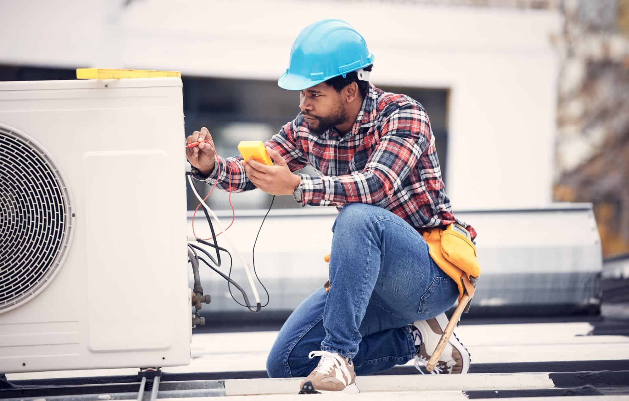 A technician wearing a blue hard hat and plaid shirt from an HVAC Contractor in San Mateo County, CA uses a multimeter to check an outdoor air conditioning unit on a rooftop.