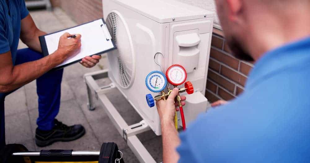 Two technicians from an HVAC Contractor in San Mateo County, CA work on an outdoor air conditioning unit; one uses a pressure gauge manifold while the other records notes on a clipboard. Tools are visible nearby.