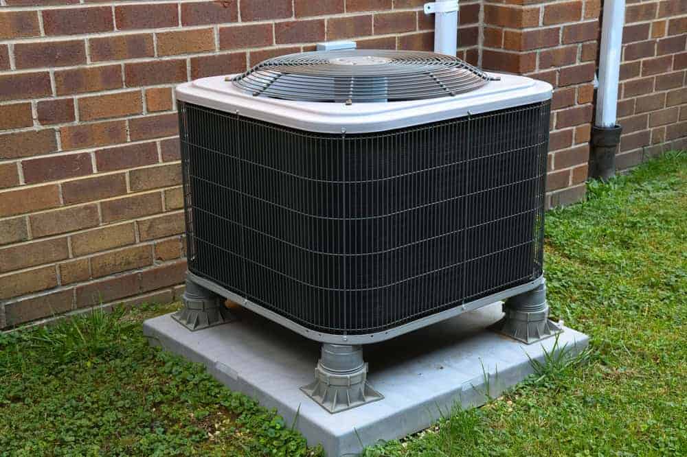 An outdoor air conditioning unit sits on a concrete slab next to a brick wall, surrounded by grass and pipes attached to the wall—professionally installed by an HVAC Contractor in San Mateo County, CA.